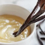A jar of homemade vanilla bean paste with visible specks of vanilla seeds, placed on a wooden counter next to vanilla pods and a small bowl of sugar.