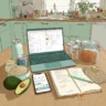 Illustration of a cozy kitchen table with a laptop showing a recipe planner, a smartphone, an open recipe notebook, jars of grains, and fresh ingredients like avocado.