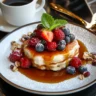 3 ingredient oatmeal pancakes topped with berries and drizzle of honey, and a cup of coffee nearby.