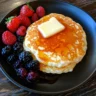 Stack of golden sweet cream pancakes topped with melting butter and maple syrup, surrounded by fresh berries.