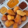 A plate of golden, crispy chicken nuggets with dipping sauces on a rustic wooden table.
