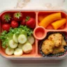 A colorful lunch box divided into compartments, each filled with kid-friendly foods such as sliced apples, baby carrots, cheese cubes, crackers, a small sandwich shaped like a star, and a container of hummus, arranged neatly to appeal to picky eaters.