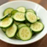 A bowl of fresh cucumber salad with thinly sliced cucumbers, red onions, and dill, lightly dressed in vinegar and oil, served on a wooden table with a fork on the side.
