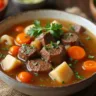 A steaming bowl of beef broth soup filled with tender chunks of beef, diced carrots, celery, and onions, garnished with fresh parsley and served in a white ceramic bowl on a rustic wooden table.