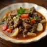 A bowl of beef sinigang filled with tender beef chunks, vibrant vegetables like okra, eggplant, string beans, and water spinach, all in a steaming, tangy tamarind-based broth, served on a rustic wooden table.