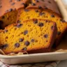 A freshly baked loaf of chocolate chip pumpkin bread sliced to reveal a moist, orange-hued interior studded with melted chocolate chips, sitting on a wooden cutting board with scattered chocolate chips nearby.