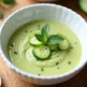 Chilled cucumber gazpacho served in a glass bowl, garnished with a drizzle of olive oil, fresh herbs, and diced vegetables, placed on a light wooden surface with a spoon beside it.