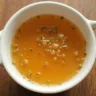 A steaming pot of homemade chicken broth filled with chicken pieces, carrots, celery, onions, and herbs like parsley and thyme simmering on a stovetop with a ladle resting nearby.