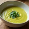 Bowl of leek soup on a wooden table, garnished with fresh herbs, surrounded by whole leeks, carrots, and celery, promoting a healthy, low-calorie option for weight loss