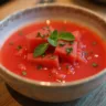 A bowl of chilled watermelon gazpacho garnished with diced cucumbers, fresh herbs, and a drizzle of olive oil, served on a rustic wooden table with slices of watermelon in the background